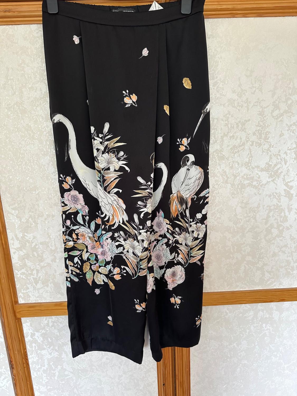 Front view of black trousers with a print of cranes and flowers in pastel shades.