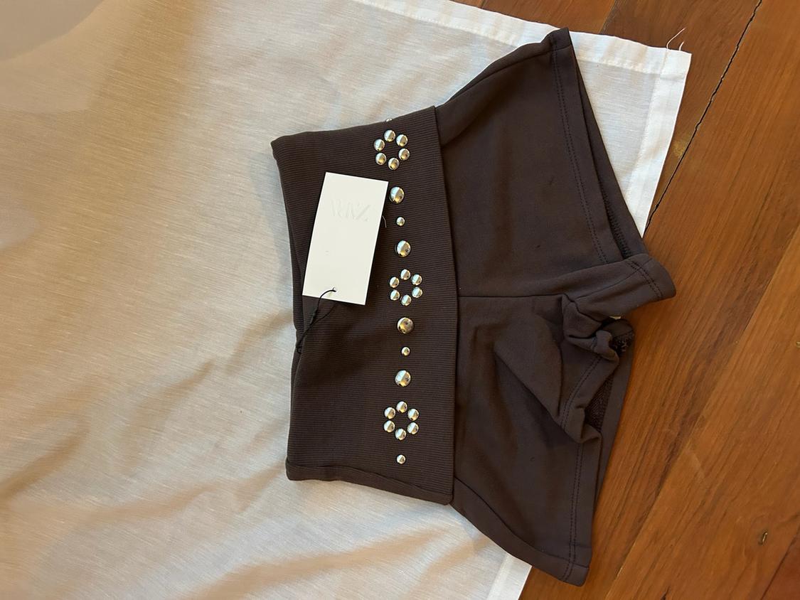 Brown shorts with a folded waistband and silver flower-shaped studs, on white fabric and wood. ZARA label.