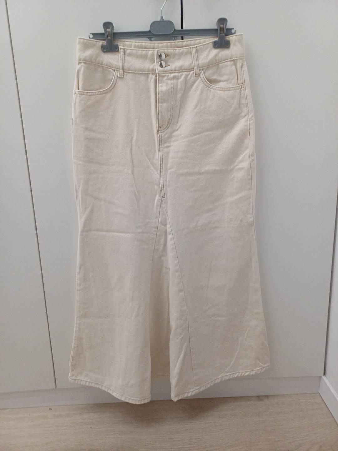 Long beige skirt hanging on a hanger in front of a white wardrobe. It features a panelled design, front pockets, and a button fastening.