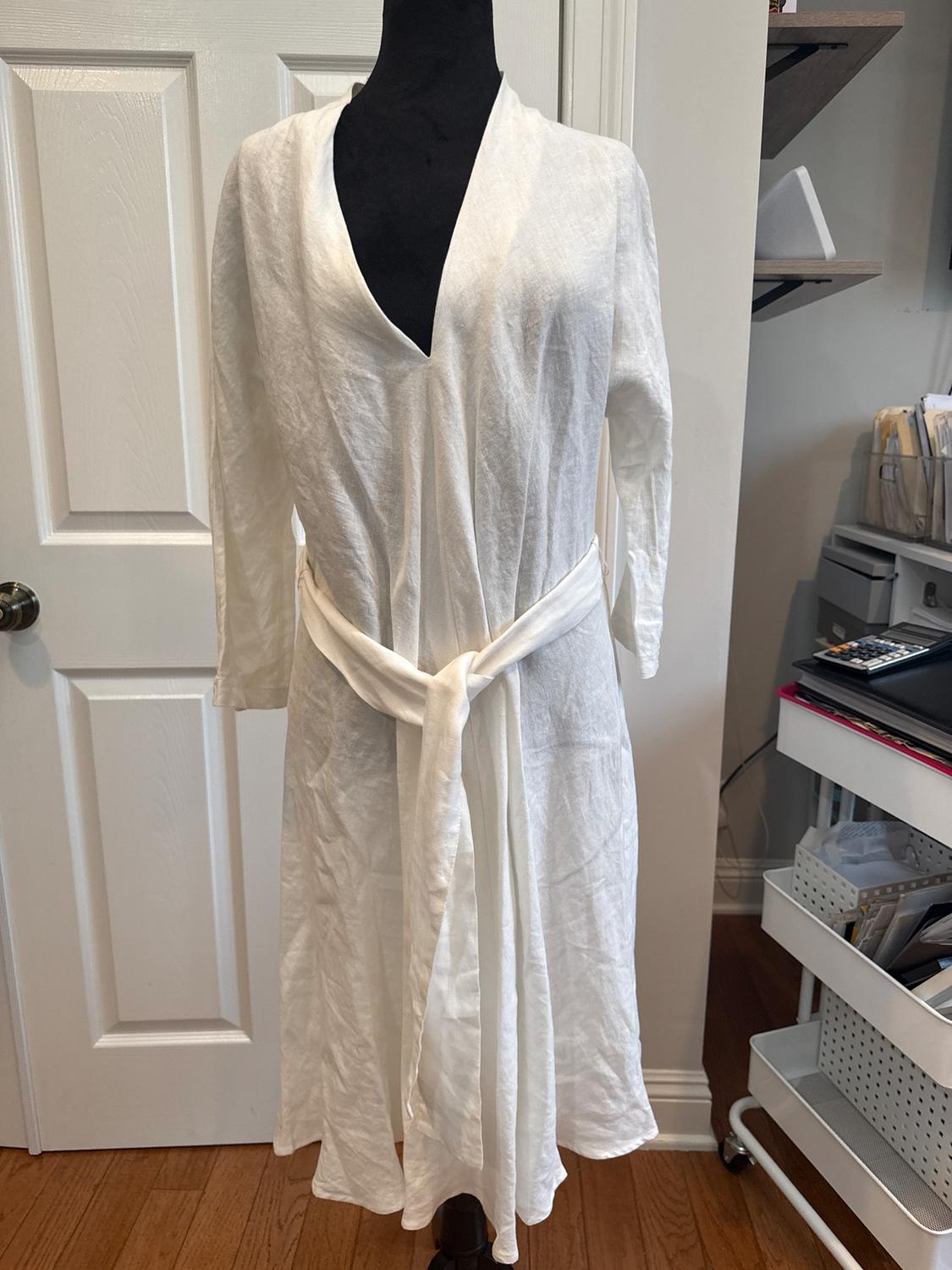 Mannequin in a white linen dressing gown, with long sleeves and a tied belt, in a room with a white door and a bookshelf in the background.