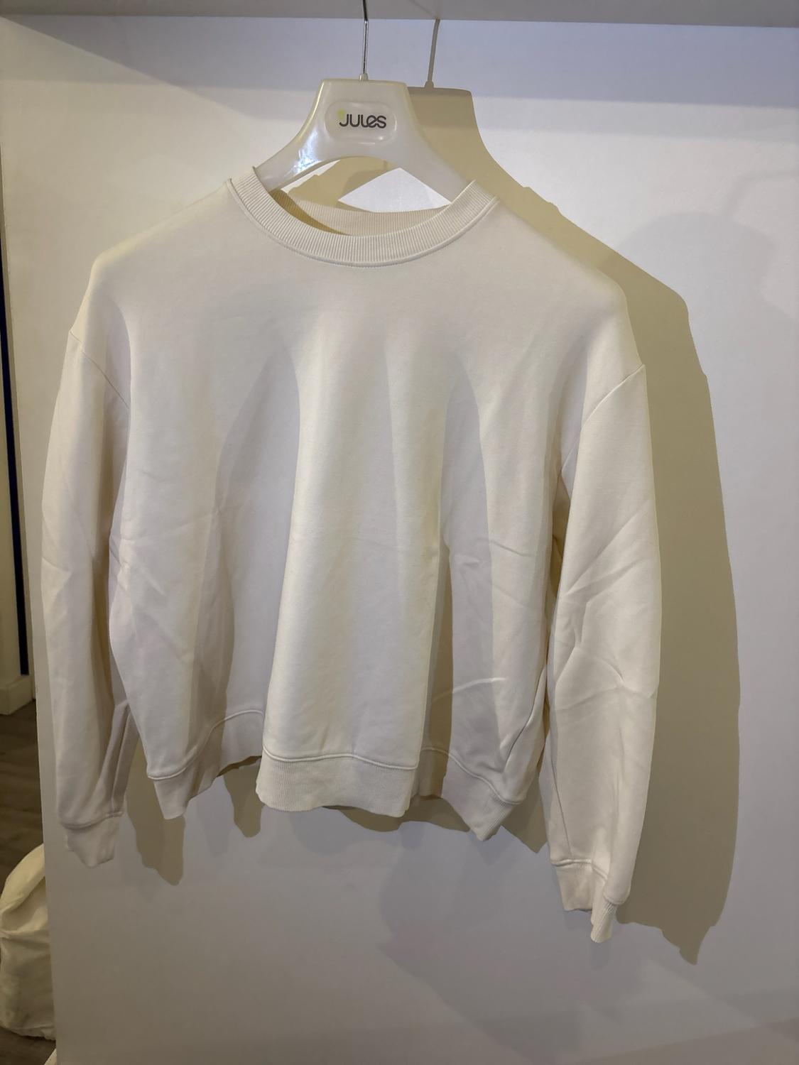White long-sleeved sweatshirt hanging on a hanger with the text JULES, casting a shadow on a light-coloured wall.
