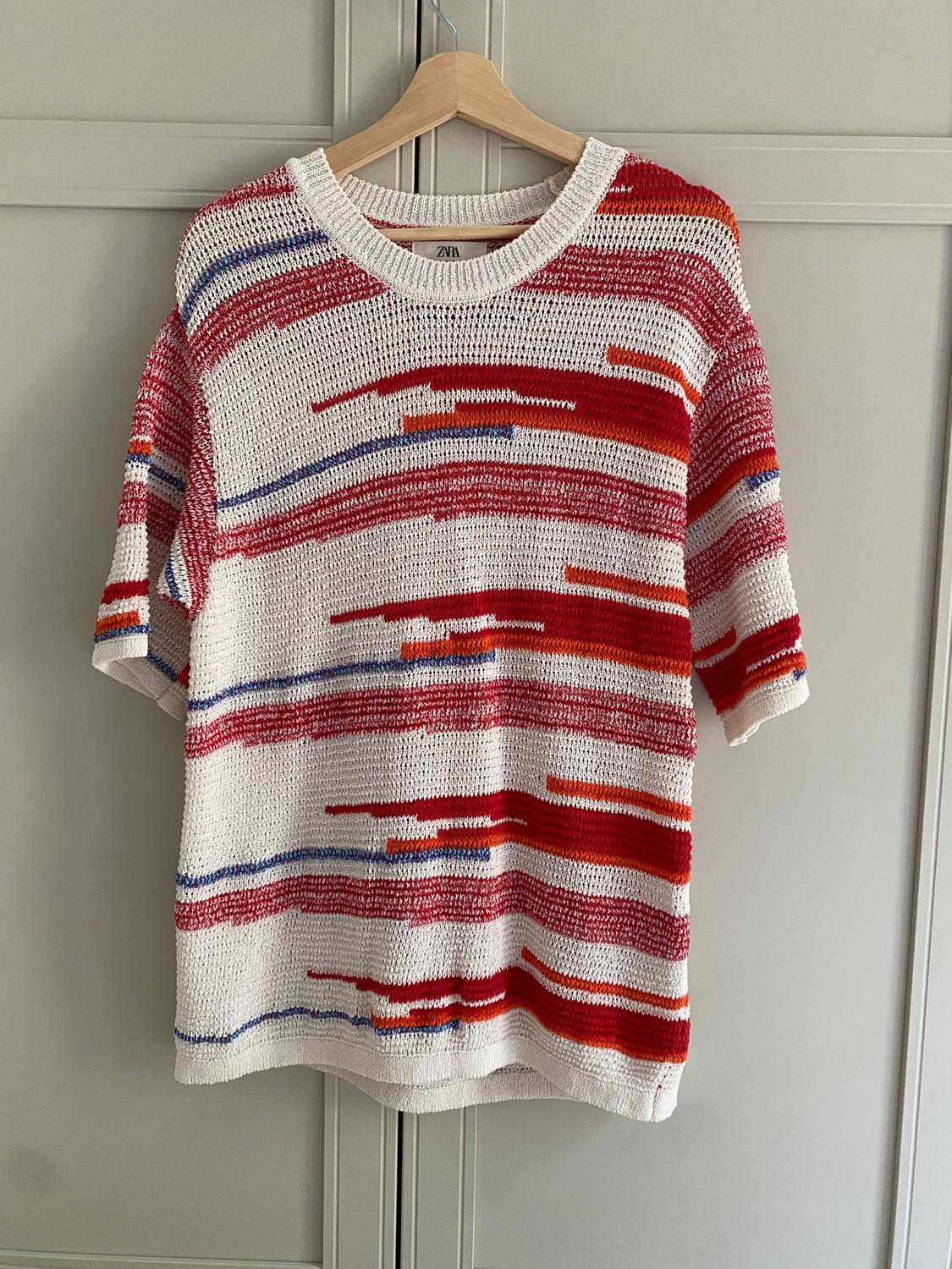 Knitted jumper hanging on a hanger, with horizontal red, blue, and orange stripes on a white background. Visible label: Zara.
