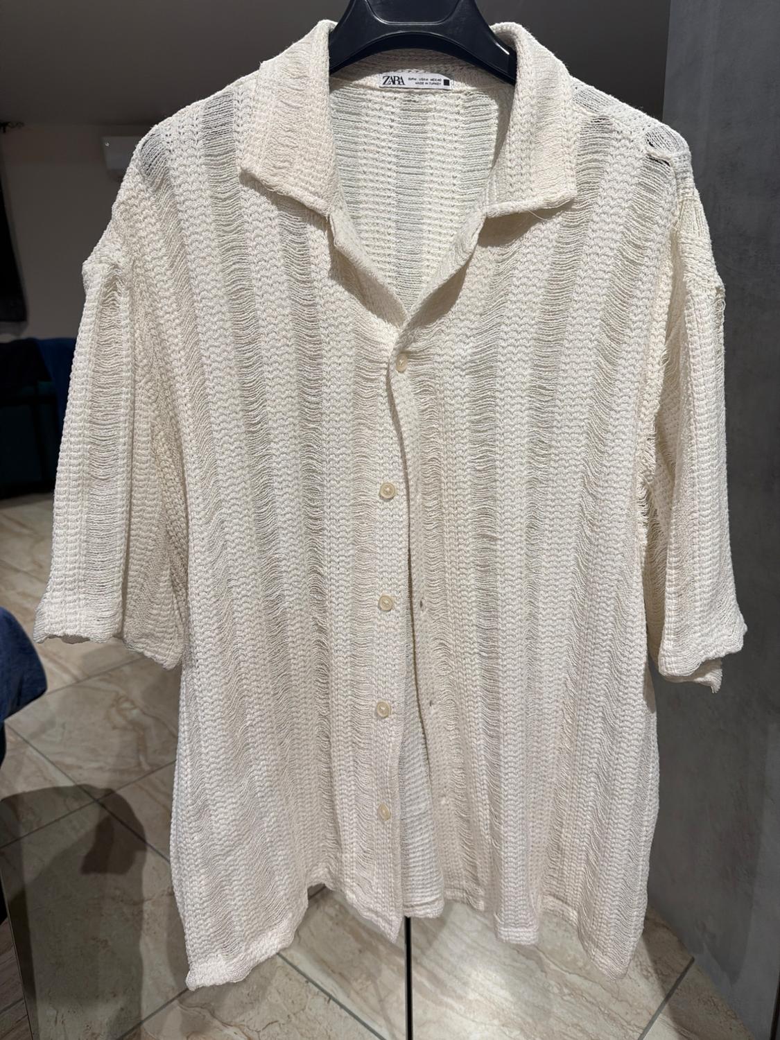 White knitted shirt with ribbed texture, short sleeves, and front buttons, hanging on a black hanger against a neutral background.