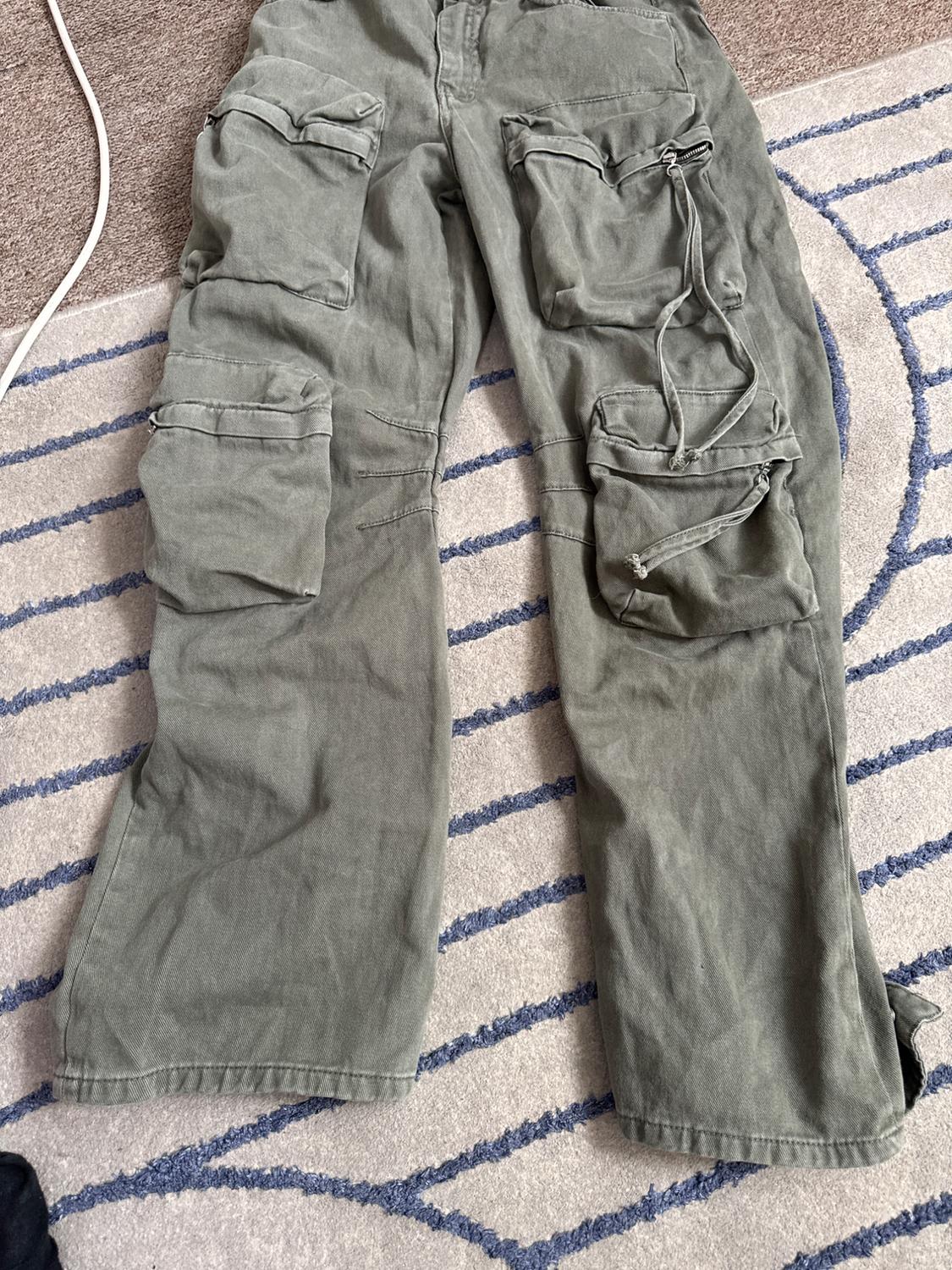 Olive green cargo trousers with large pockets and hanging straps, laid out on a light rug with wavy blue lines.