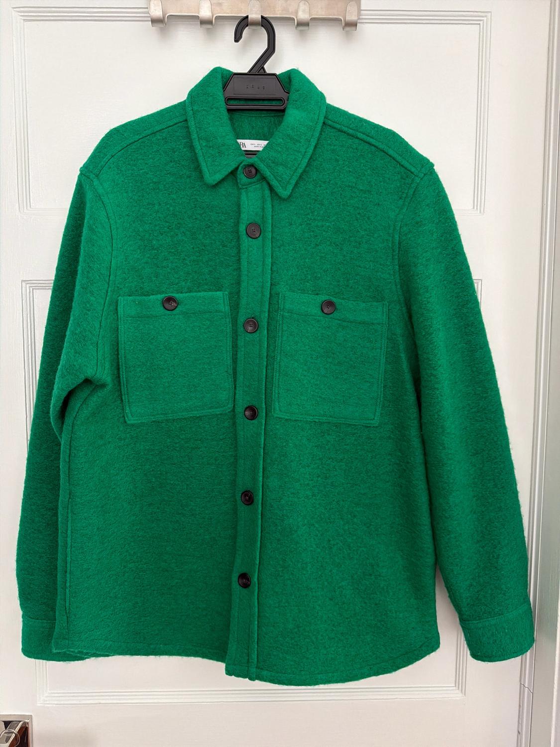 Textured green overshirt with dark buttons and two chest pockets, hanging on a black hanger over a white door.