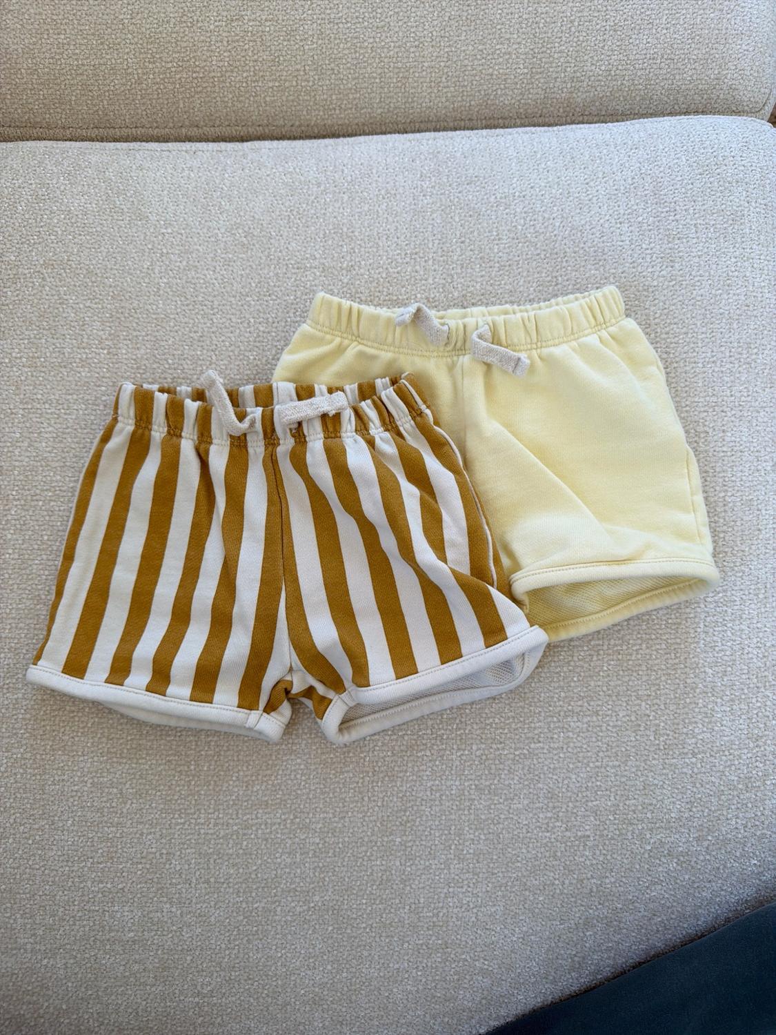 Two kids' shorts on a textured surface: one with vertical mustard and white stripes, and the other in solid yellow.