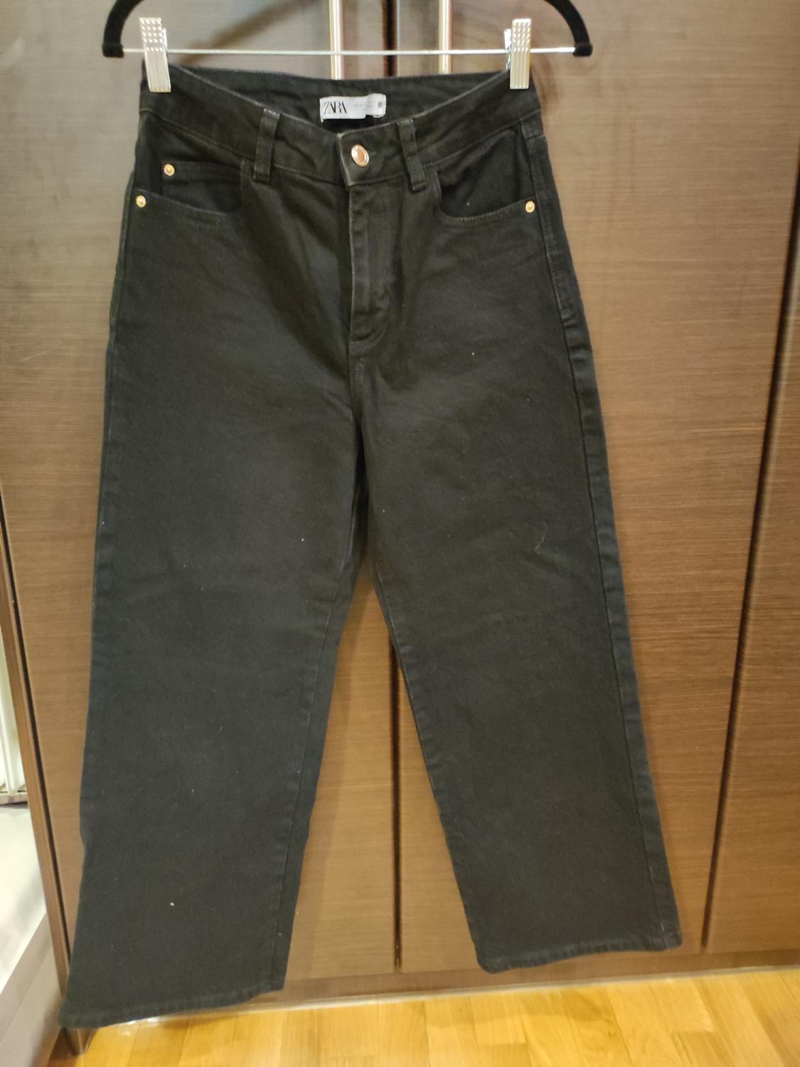 Black trousers hanging on a hanger in front of a wooden wardrobe. Visible label with the text "ZARA".