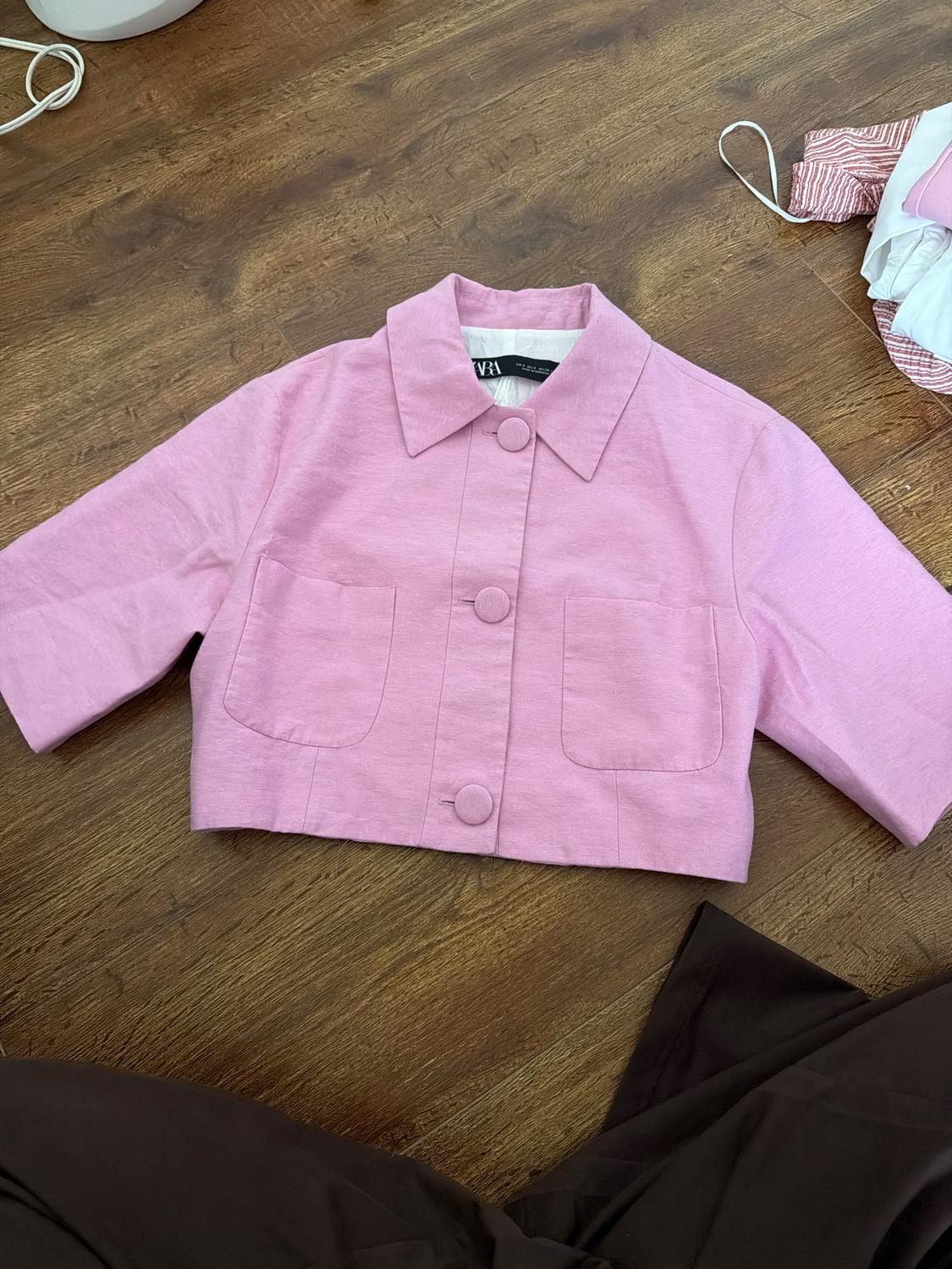Pink short jacket with large buttons and front pockets on a wooden floor. Part of dark trousers visible in the corner.