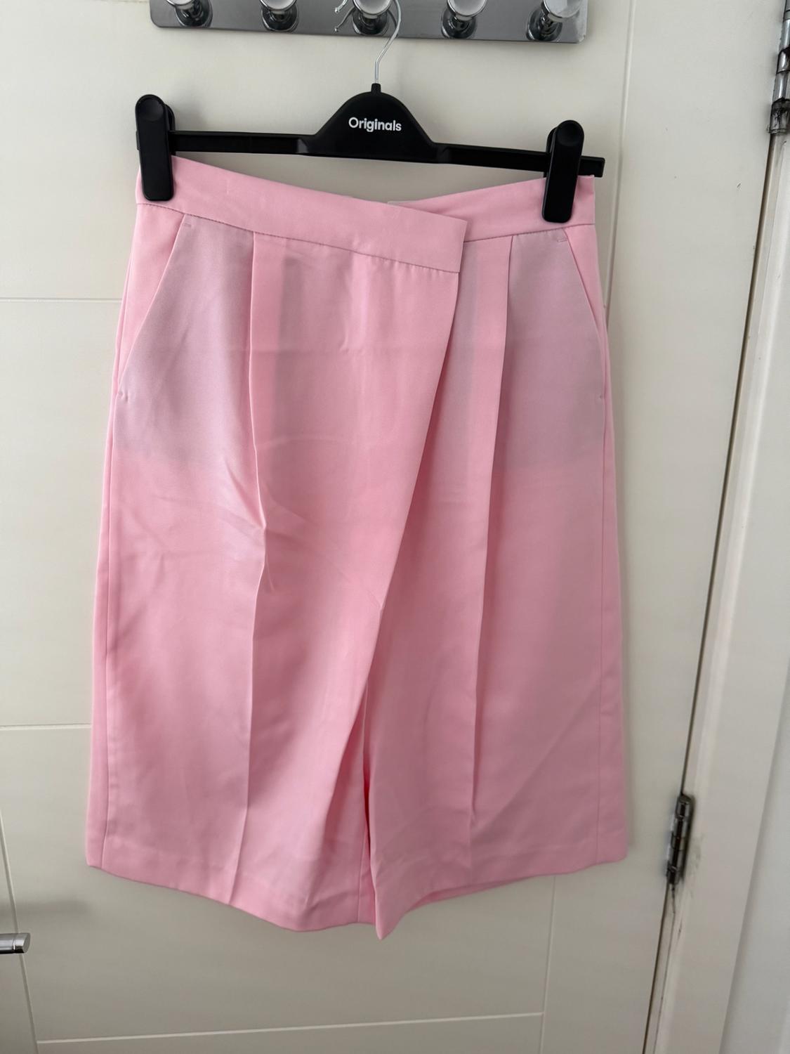 Light pink shorts with a crossed design hanging on a black hanger, with the text ‘Originals’, on a white door.