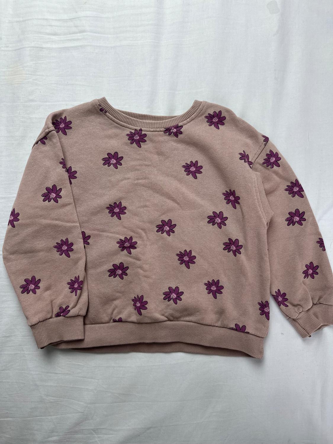 Beige sweatshirt with a purple floral print, a crew neck, and ribbed cuffs, on a white fabric background.