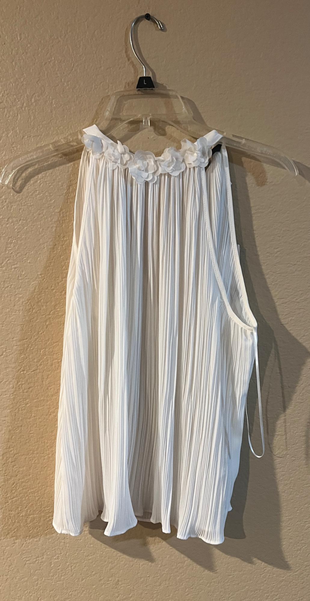 White pleated blouse with floral detailing at the neck, hanging on a transparent hanger against a beige wall.