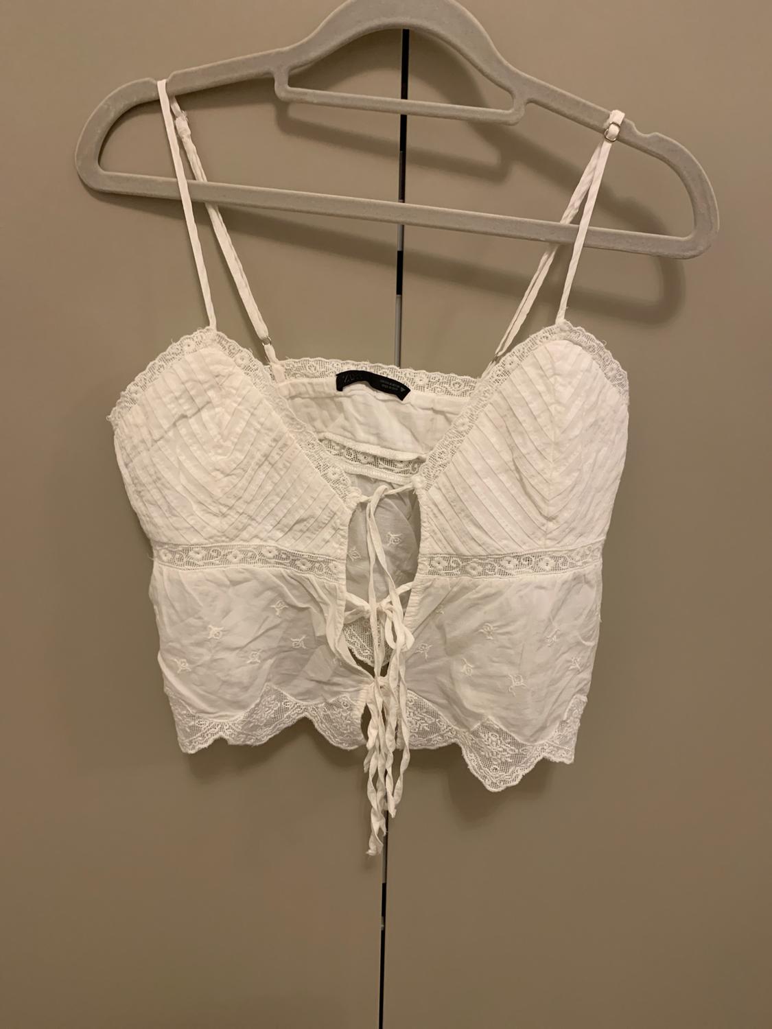 White camisole top with lace and front lacing, hanging on a grey hanger against a beige background.