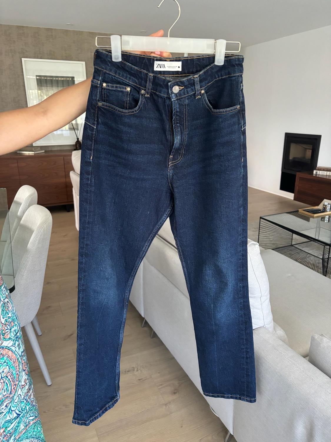 Arm holding a pair of dark blue jeans with a ZARA label on a hanger. In the background, a modern living room with a white sofa.
