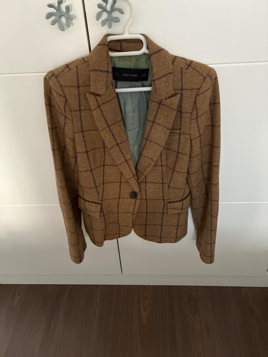 Brown checked blazer hanging in a white wardrobe. Interior label with the text ZARA BASIC. Wooden floor visible.