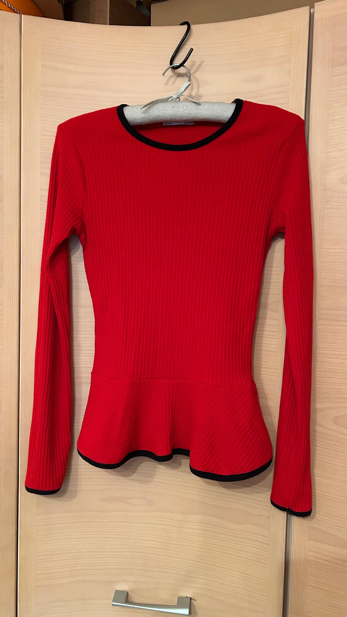 Red long-sleeved ribbed jumper with black trim, hanging on a hanger over a light wooden door.