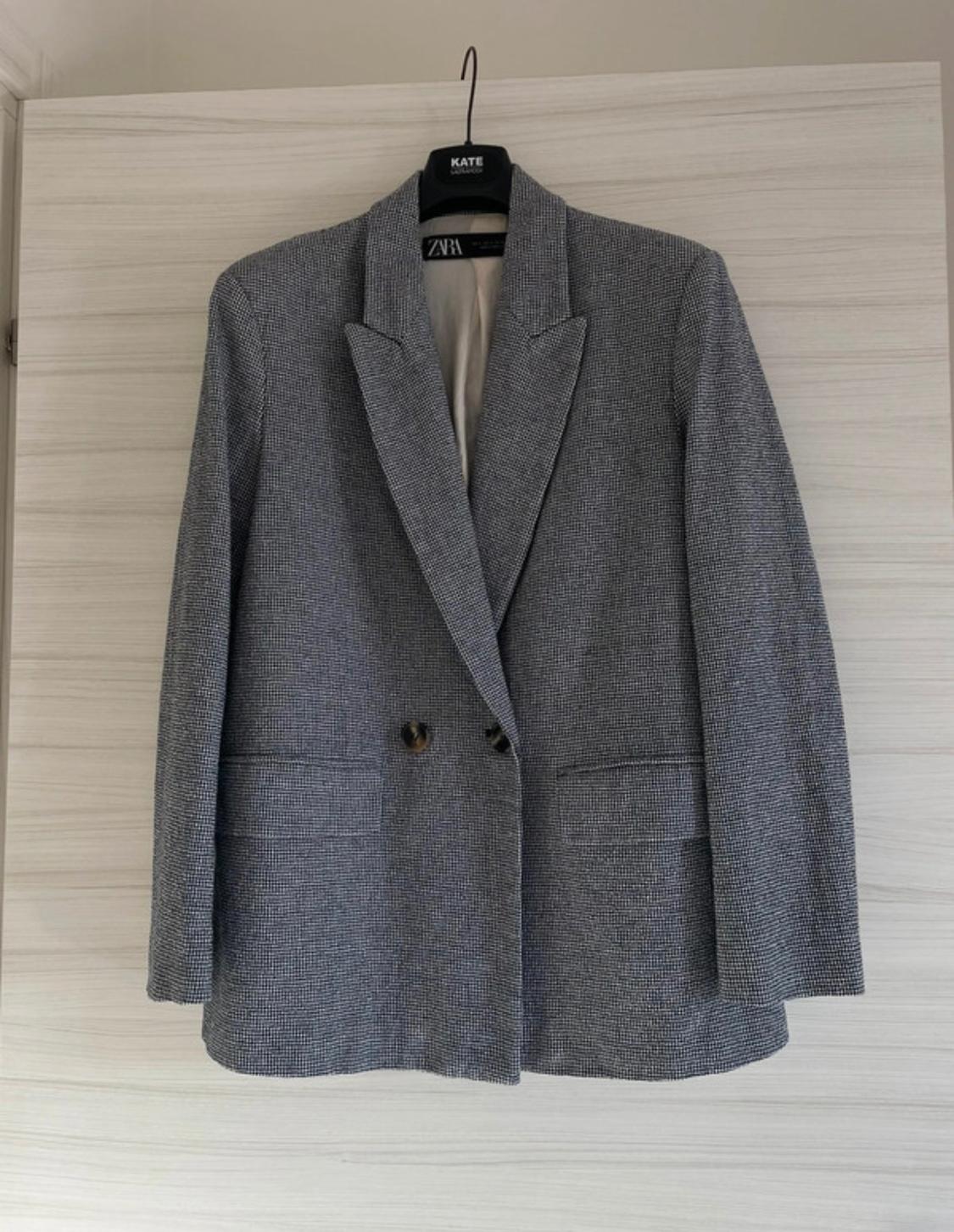 Two-button grey blazer hanging on a black hanger with a visible "ZARA" label, against a light wood background.