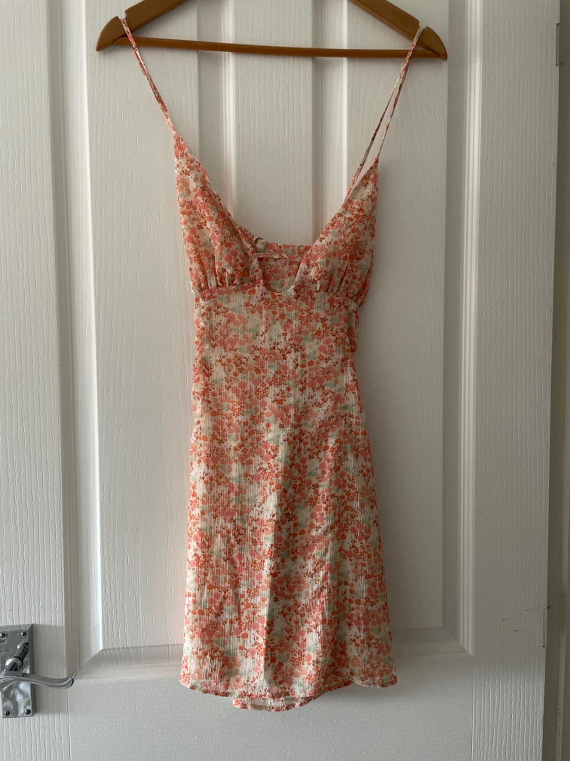 Short, thin-strapped dress with an orange and green floral print, hanging on a wooden hanger over a white door.
