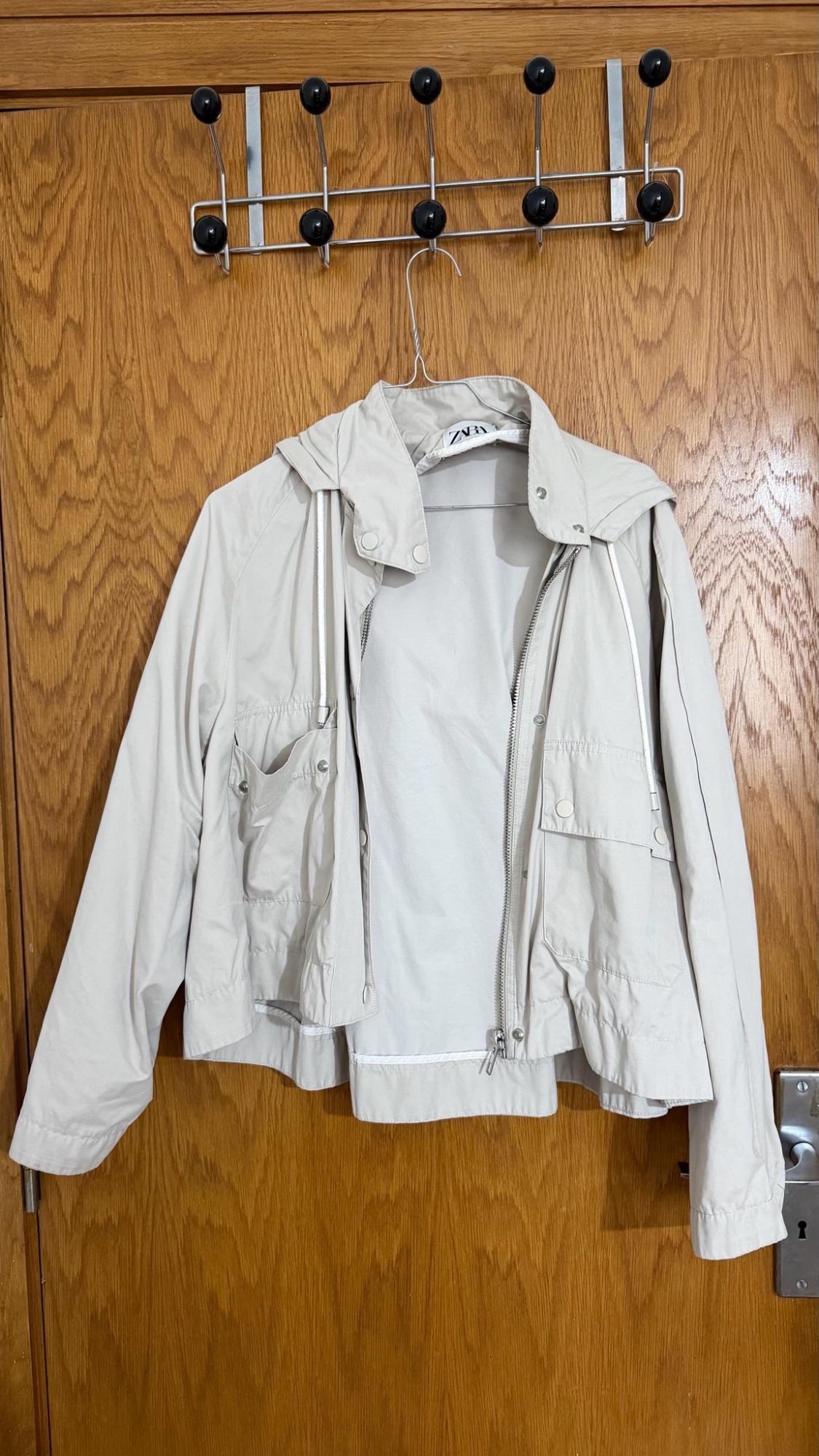 Beige hooded jacket with pockets, hanging on a hanger over a wooden door. ZARA label.