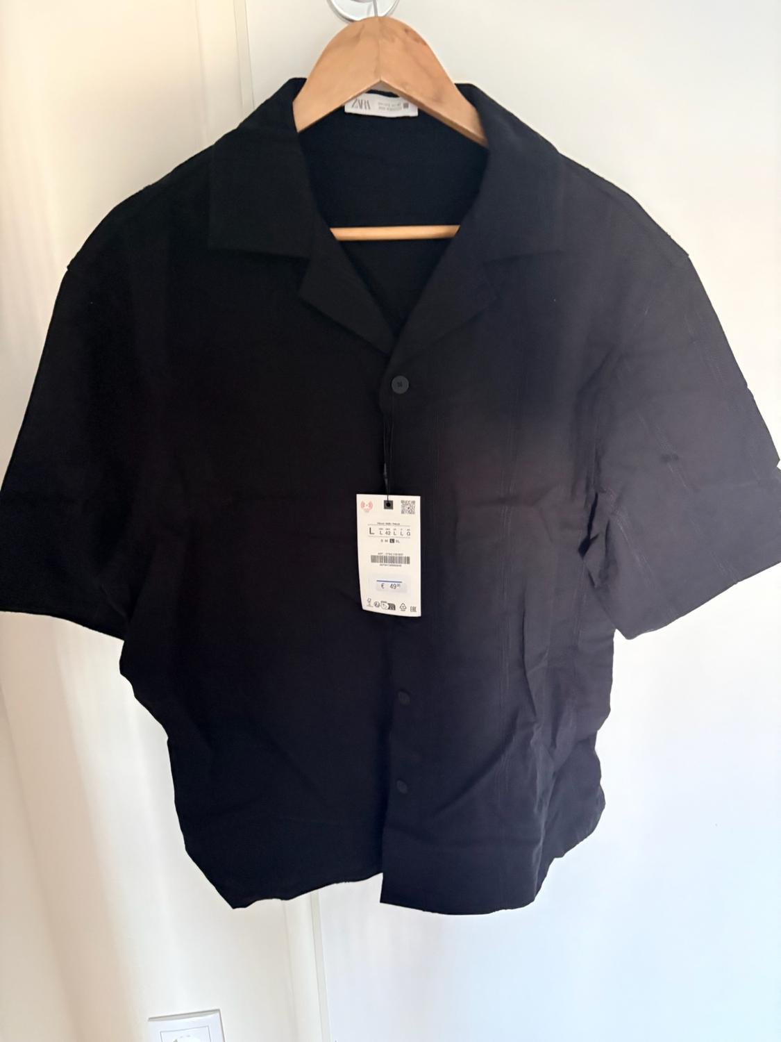 Black short-sleeved button-up shirt hanging on a wooden hanger against a white background. Neck label with the text ZARA.