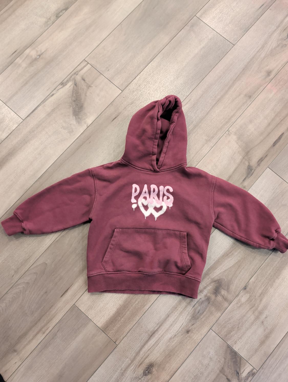 Burgundy hoodie on a light wooden floor. "PARIS" text on the chest with a heart design.