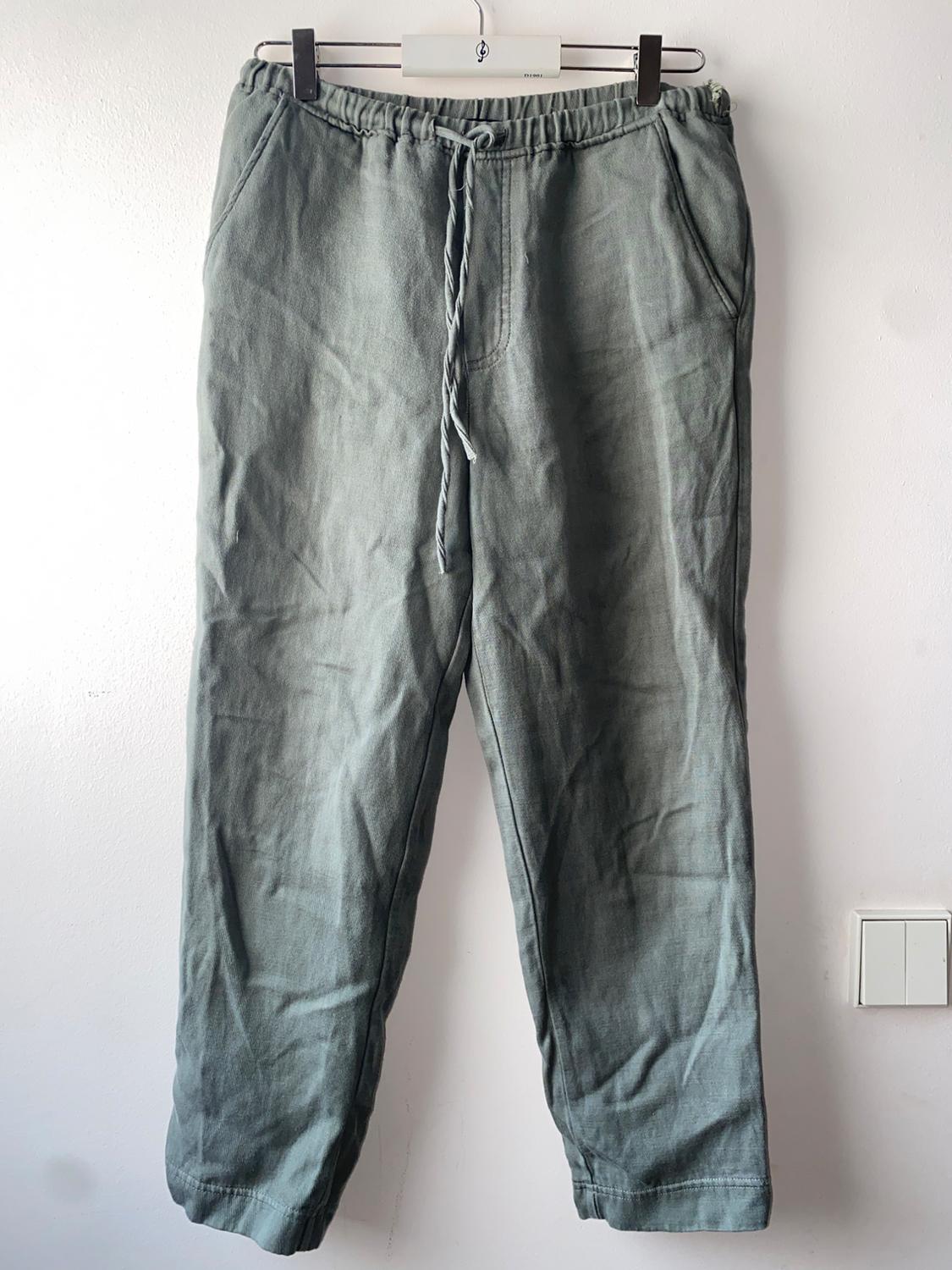 Olive green trousers in a light fabric with an adjustable drawstring waist, hanging on a metal hanger against a white wall.