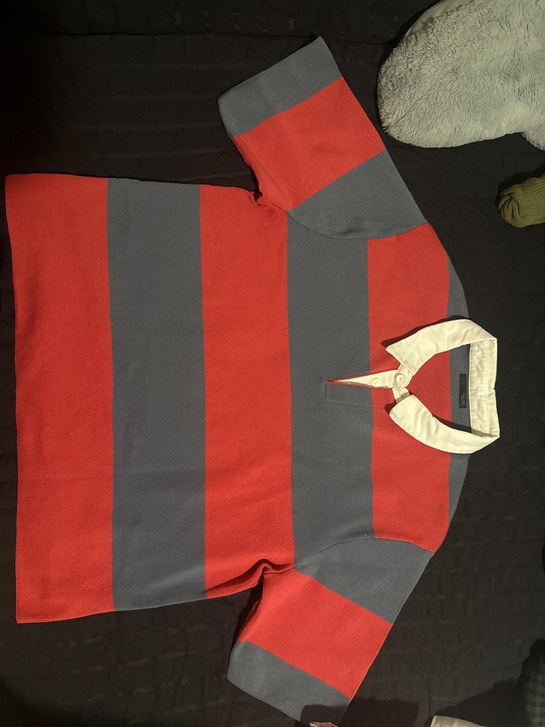 Short-sleeved polo shirt with wide red and gray stripes and a white collar. The garment is laid out on a dark surface. Casual style.