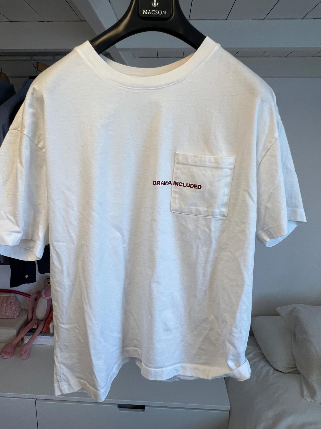 White t-shirt hanging on a black hanger with "DRAMA INCLUDED" text on the pocket. Room background with bed and toys.