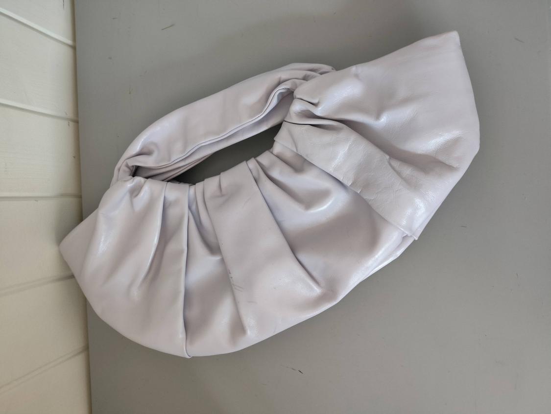 Light lilac ruched shoulder bag, resting on a grey surface next to a white wall.