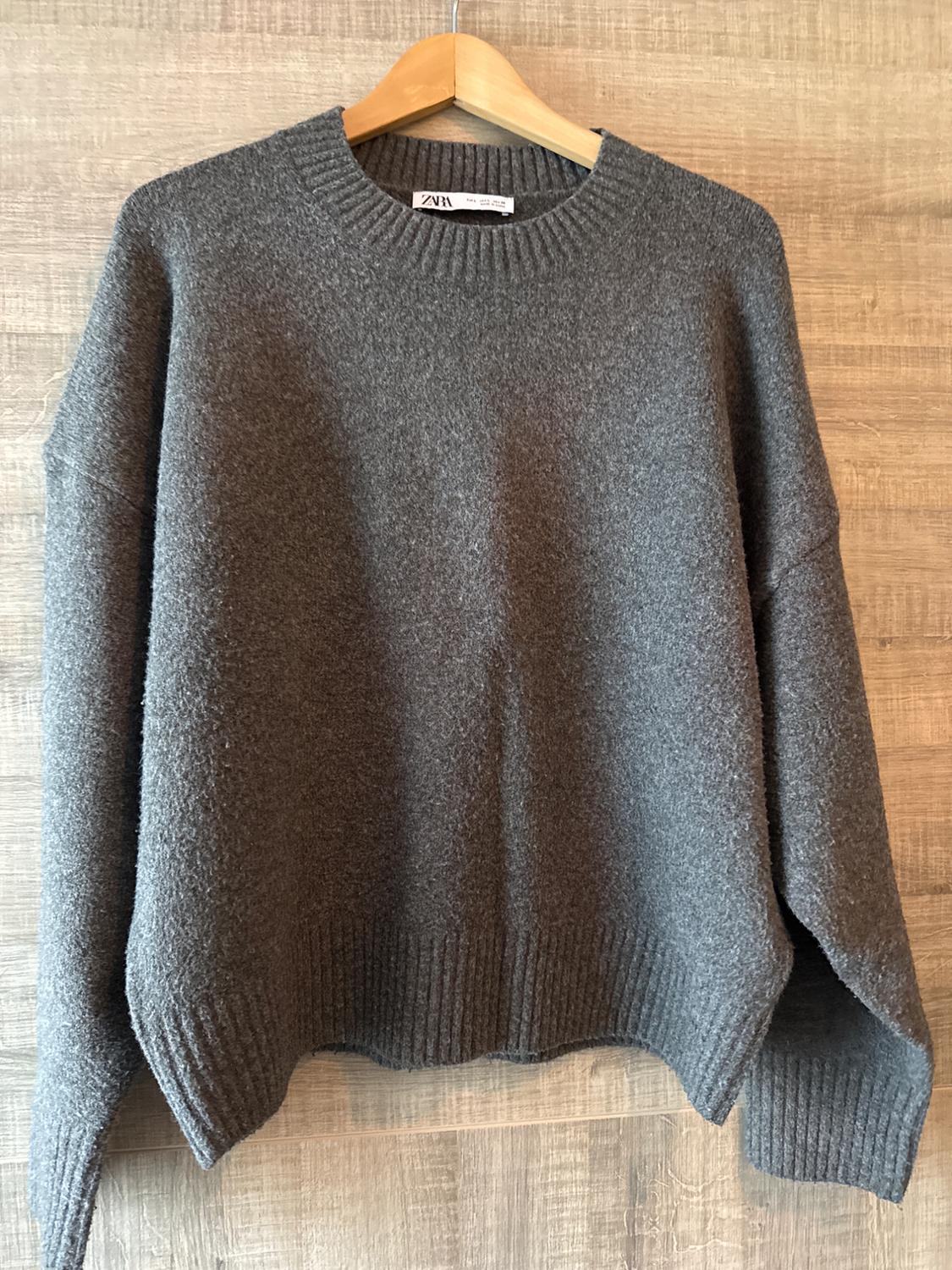 Chunky knit grey jumper hanging on a wooden hanger against a light wooden background. Label visible on the collar.