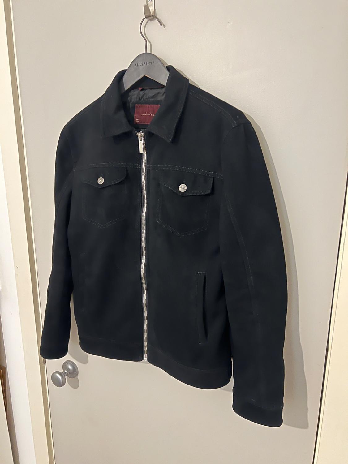 Front view of a black jacket with a zip and frontal flap pockets with buttons.