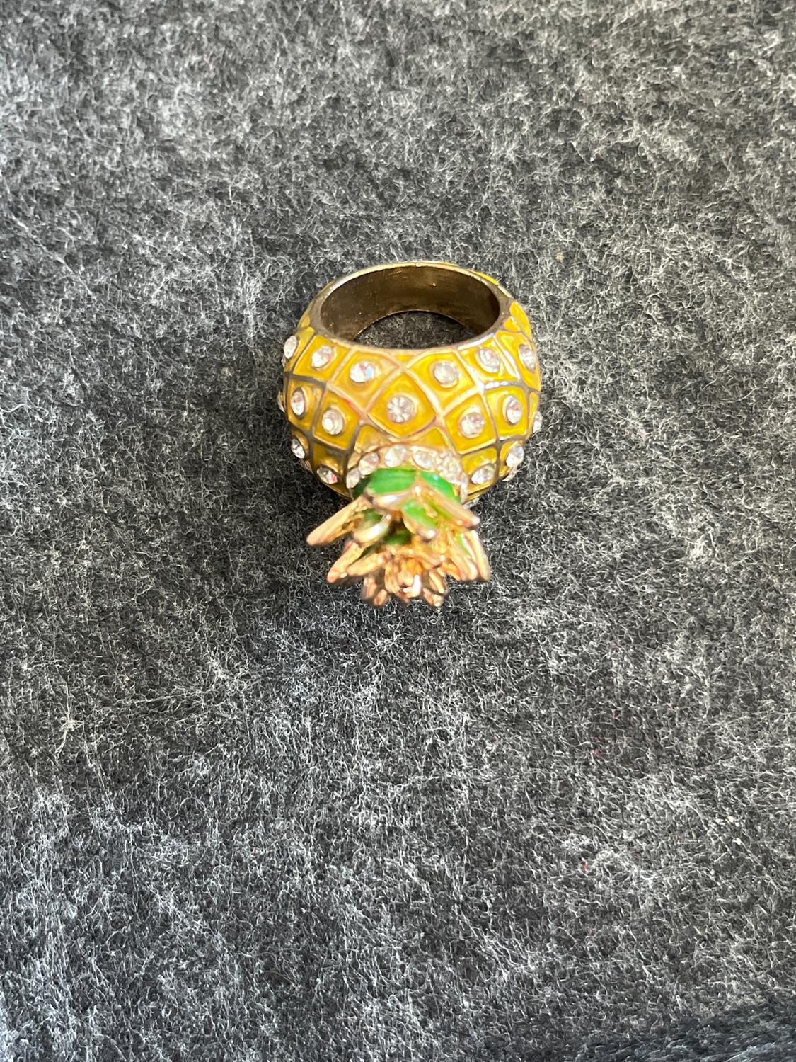 Gold ring with a pineapple design, brilliant stone details and green leaves, on a textured grey background.