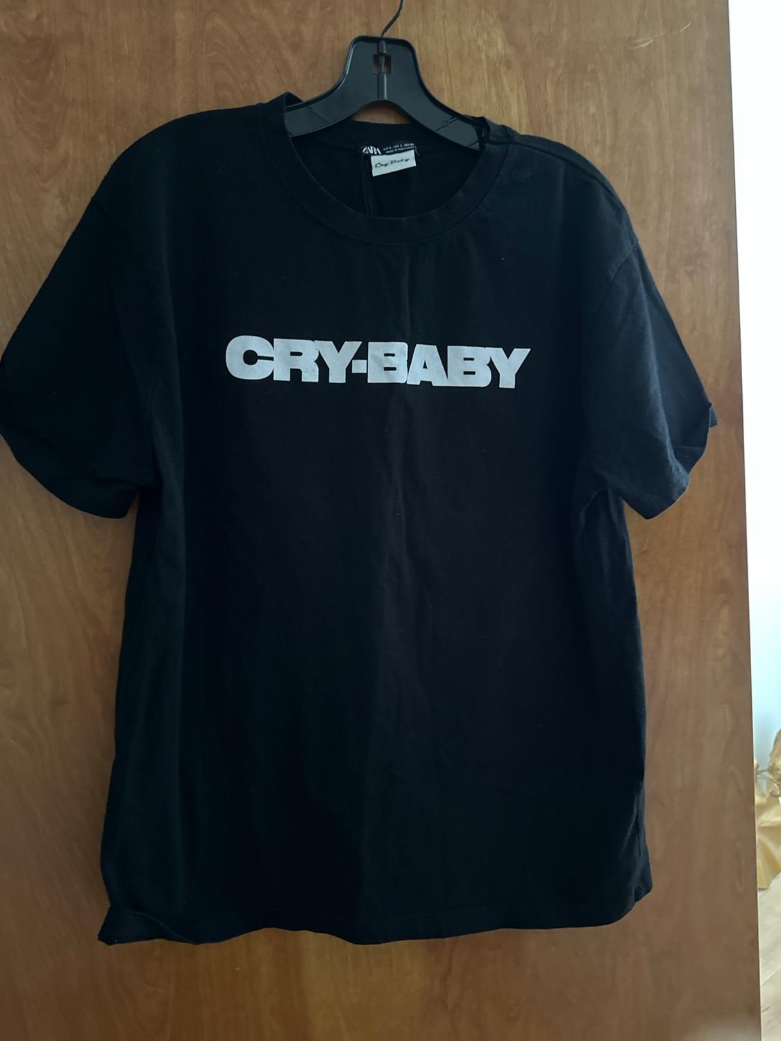 Black short-sleeved T-shirt on a hanger over a wooden door. It shows the text CRY-BABY on the chest and ZARA on the label.