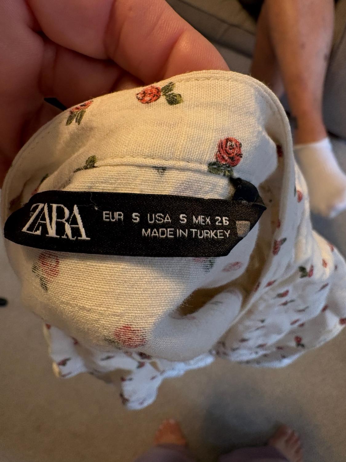Hand holding a white garment with red roses. Black label with text: ZARA EUR S USA S MEX 26 MADE IN TURKEY. Blurred background.