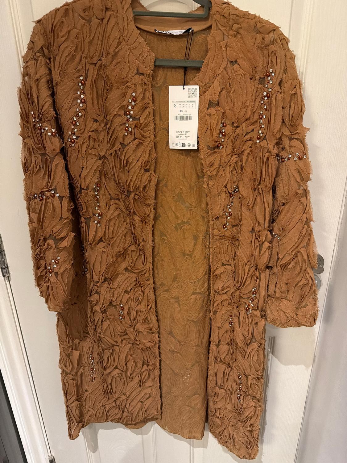 Textured brown coat with bead details, hanging on a hanger. Visible label with price and size.