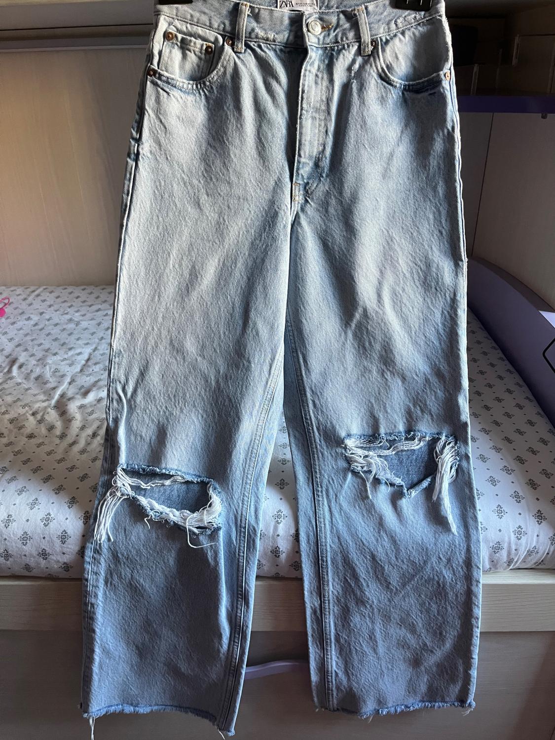 Light blue ZARA jeans with ripped knees and frayed hems, hanging from a hanger in front of a bed with patterned sheets.