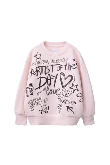 Front view of a pink sweatshirt with graphic print and the text 'ARTIST of the DAY', 'Bonjour, Friend', and others.