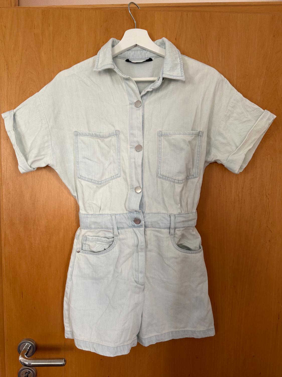 Light blue short-sleeved denim playsuit with buttons and pockets, hanging on a white hanger in front of a wooden door.