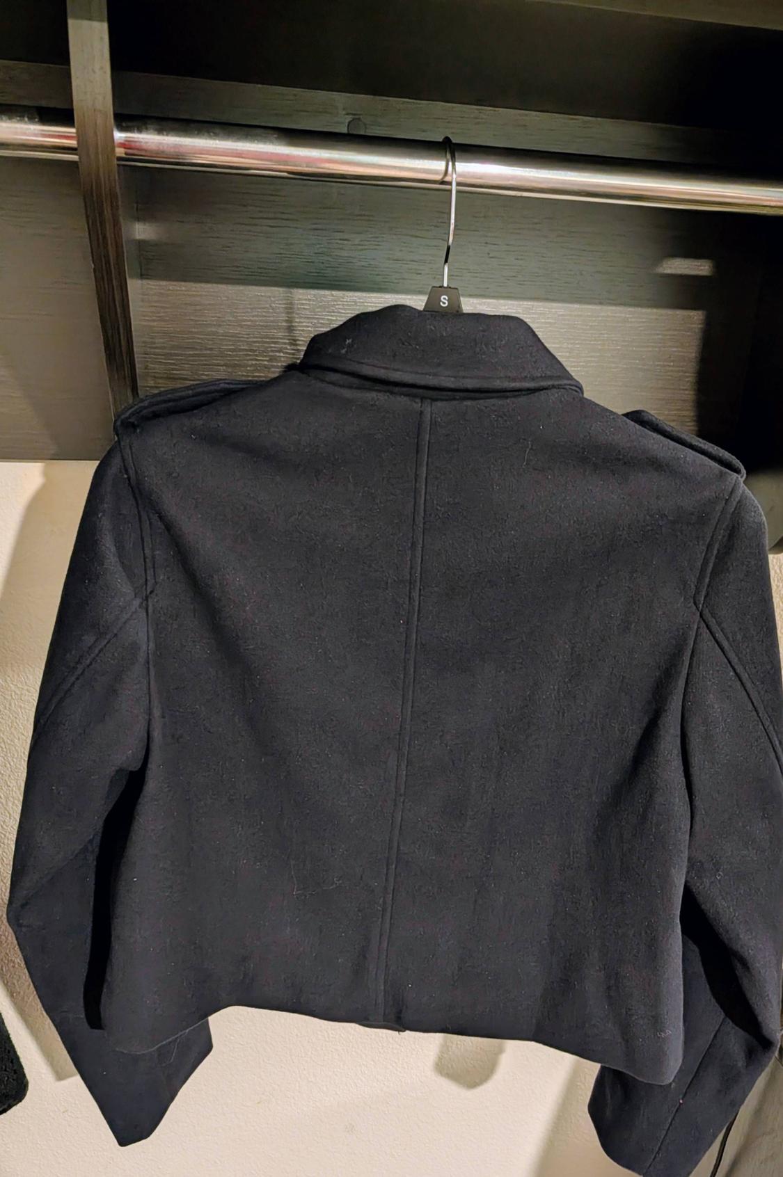 Black jacket hanging on a hanger with a size S tag, viewed from behind, in a dark wooden closet.