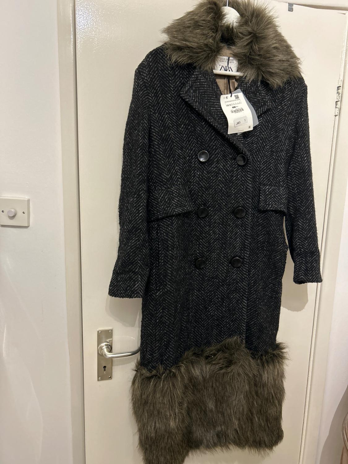 Dark grey long coat with a herringbone pattern, black buttons, and faux fur details on the collar and bottom hem, hanging on a white door.