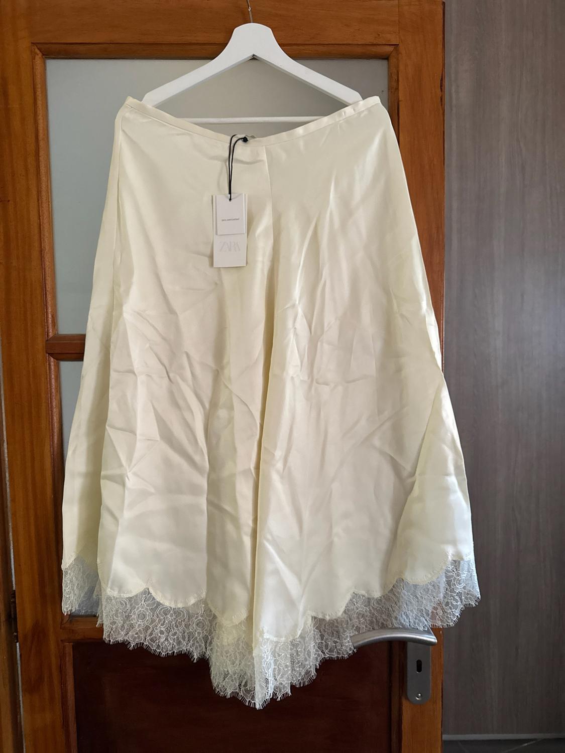Cream satin skirt with lace hem hanging on a white hanger on a wooden door. Label with the text ZARA.