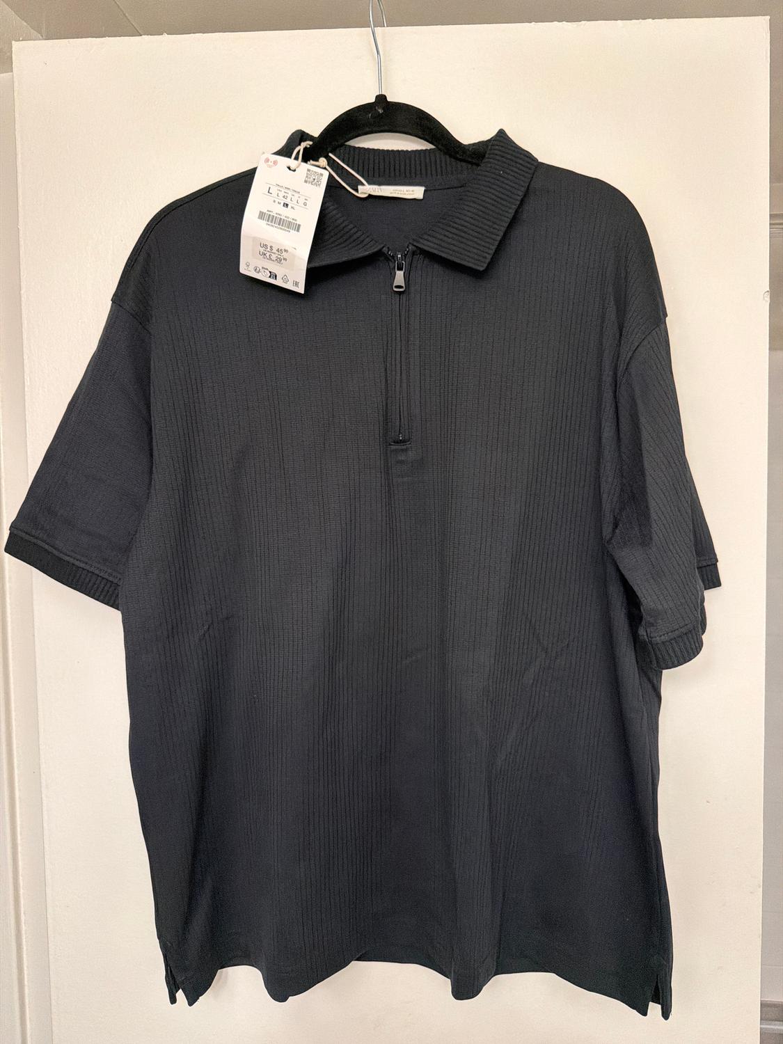 Black short-sleeved polo shirt with zip neck, hanging on a black hanger. Visible tag with price details and barcode.