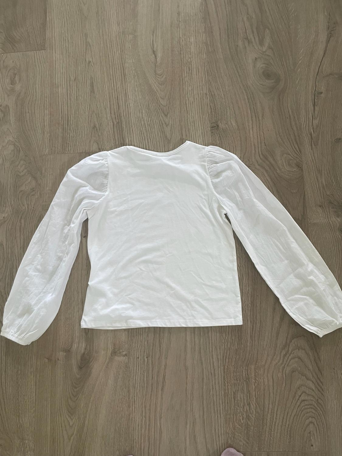 White long-sleeve T-shirt with puff shoulders, laid out on a wooden floor. Casual flat lay composition.
