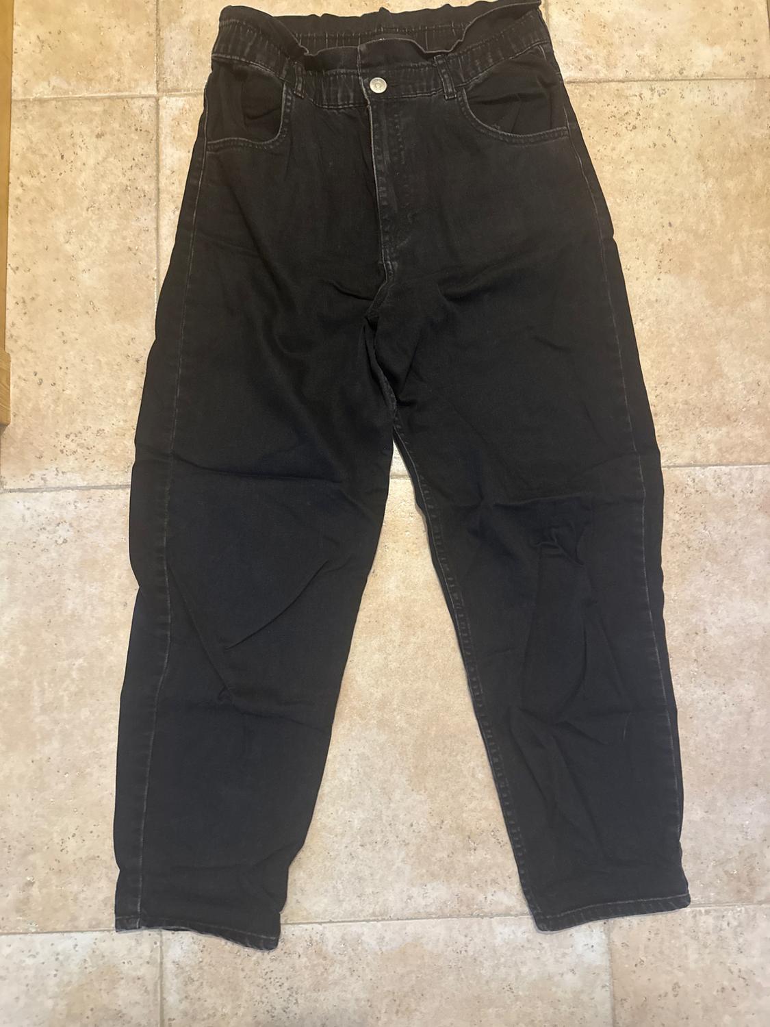 Black denim trousers with an elasticated waist and button, placed on a beige tiled floor.