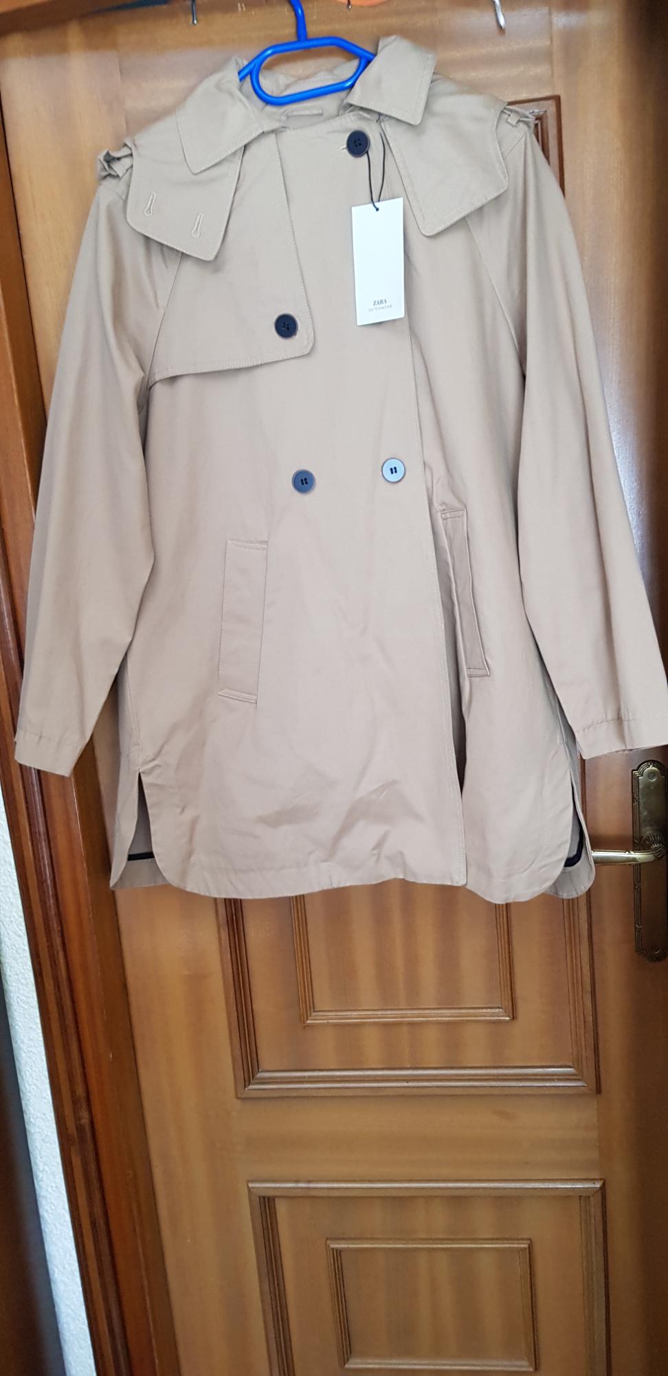 Beige coat hanging on a blue hanger over a wooden door. It has dark buttons and a white label with "ZARA" text.
