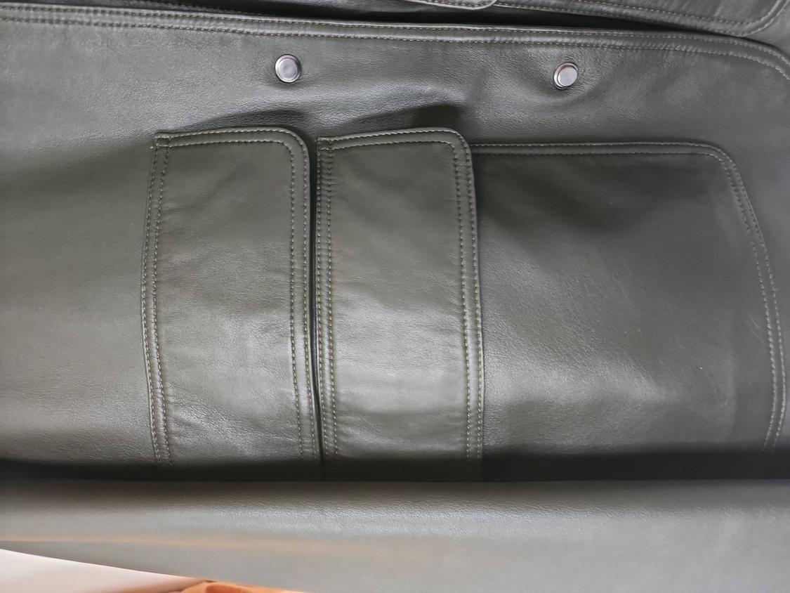 Close-up of an olive green leather garment with two flap pockets, visible stitching, and two metal buttons.