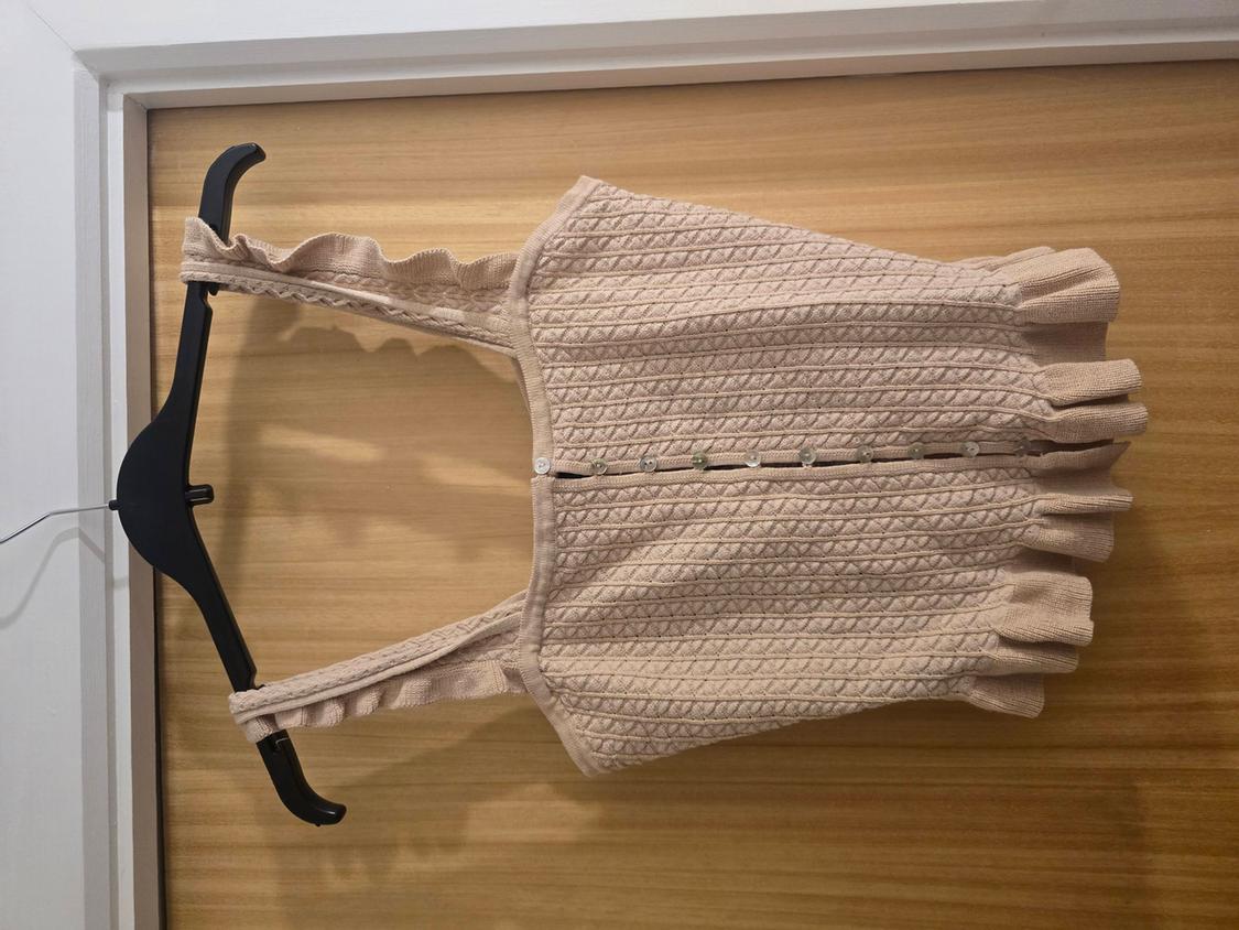 Beige knitted camisole with straps and front buttons, hanging on a black hanger against a light wood background.