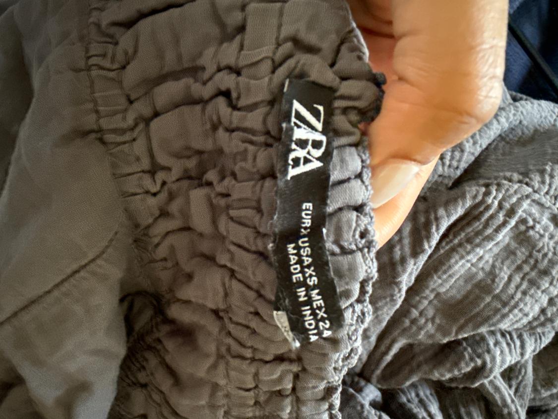Black label on wrinkled gray fabric, with the text "ZARA EUR USA XS MEX 24 MADE IN INDIA", held by a hand.