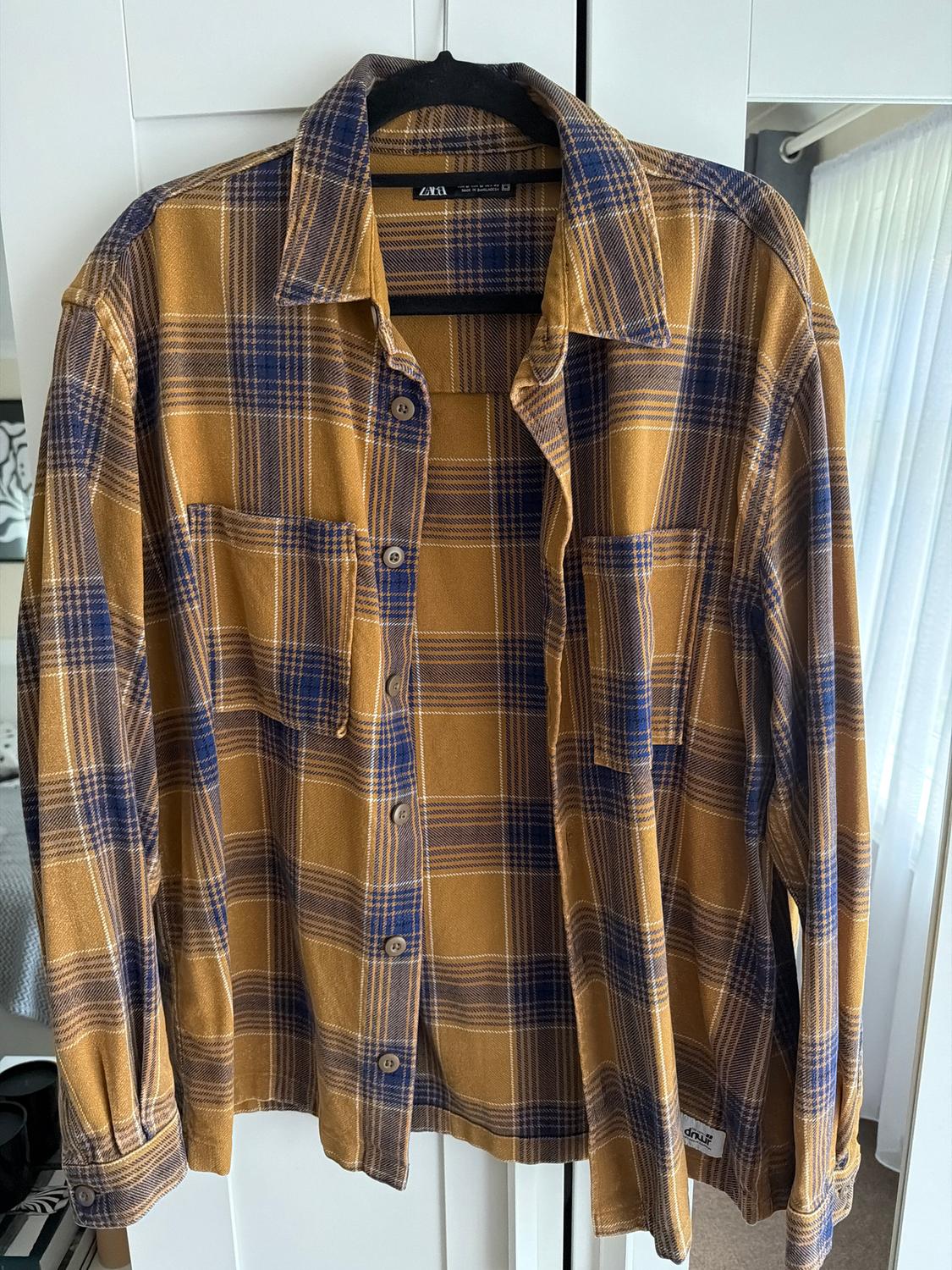 Brown and blue checked shirt hanging on a black hanger in front of a white wardrobe. Label visible: "Zara".
