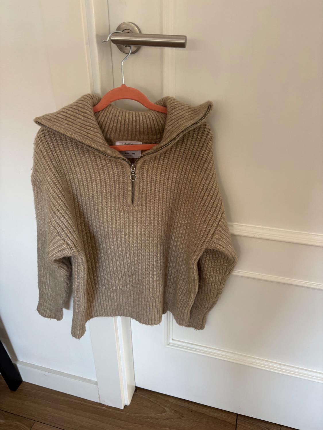 Beige knitted jumper with a zip-up neck, hanging on an orange coat hanger over a white door with a silver handle.