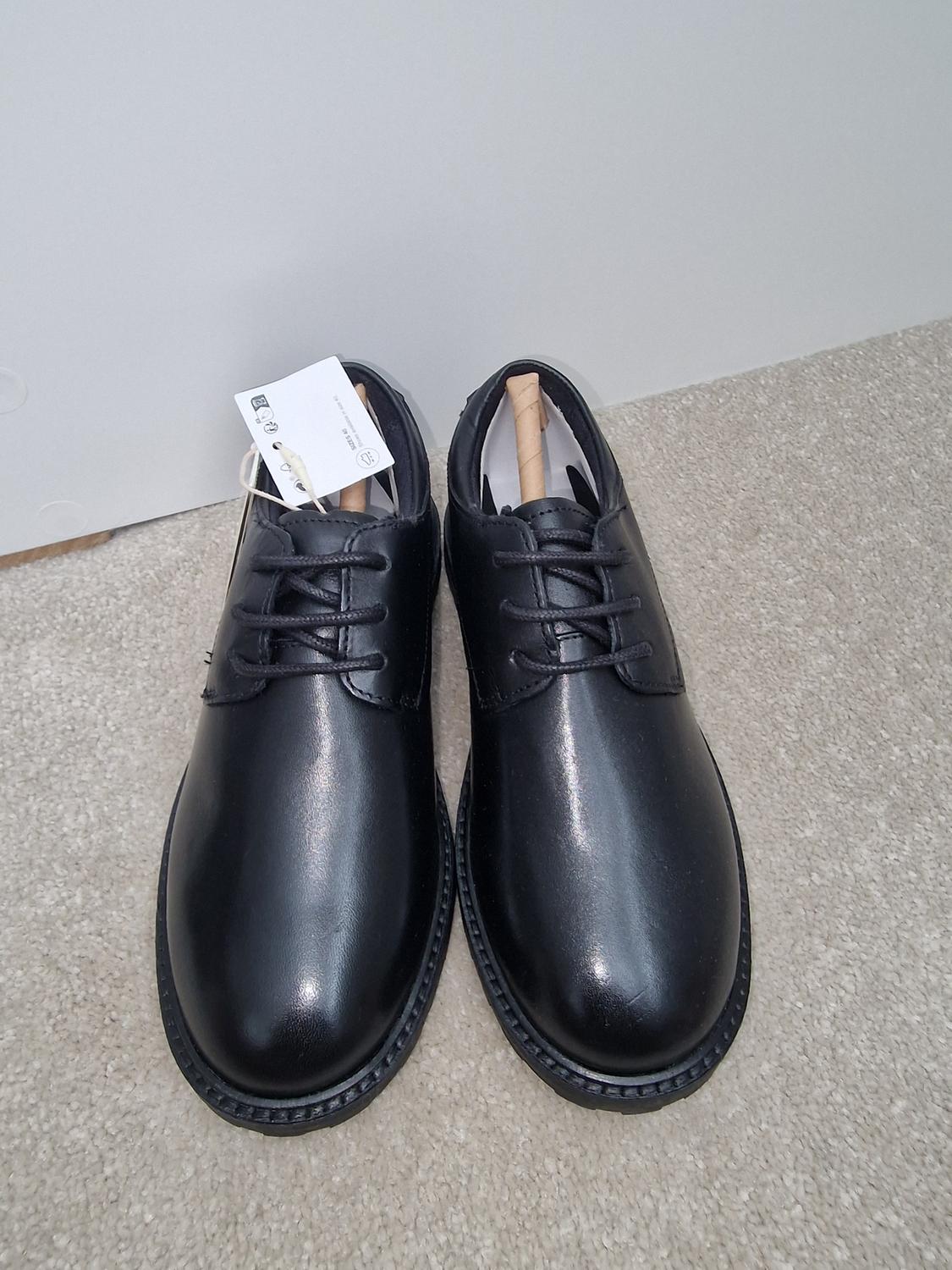 Black leather lace-up shoes on a beige carpet. White label visible on one of the shoes.
